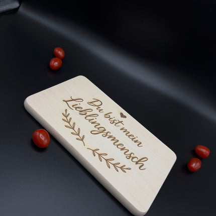 Holz-Frühstücksbrettchen mit Gravur "Du bist mein Lieblingsmensch", Herz und Blatt-Motiv auf modernem schwarzen Hintergrund.
