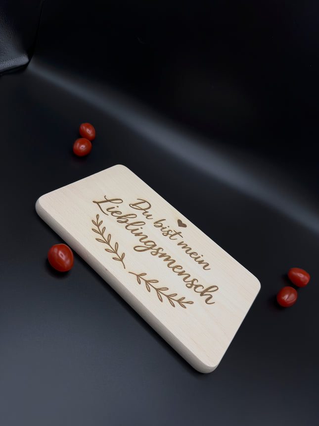 Holz-Frühstücksbrettchen mit Gravur "Du bist mein Lieblingsmensch", Herz und Blatt-Motiv auf modernem schwarzen Hintergrund.